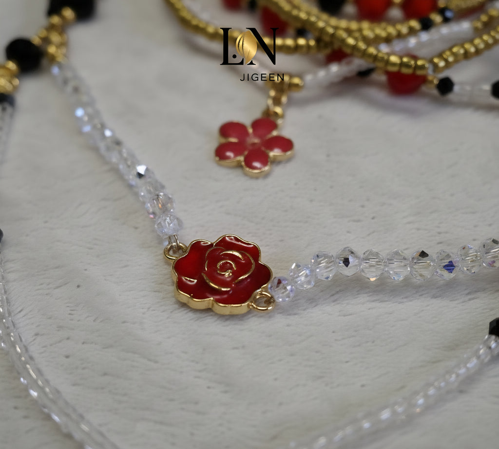 Gros plan très serré sur les perles de hanche LN JIGEEN. Détail de la ligne de perles de cristal transparentes taillées, encadrant une breloque en forme de rose émaillée rouge et or. Ce zoom met en évidence la brillance des matériaux et la précision de l'assemblage artisanal