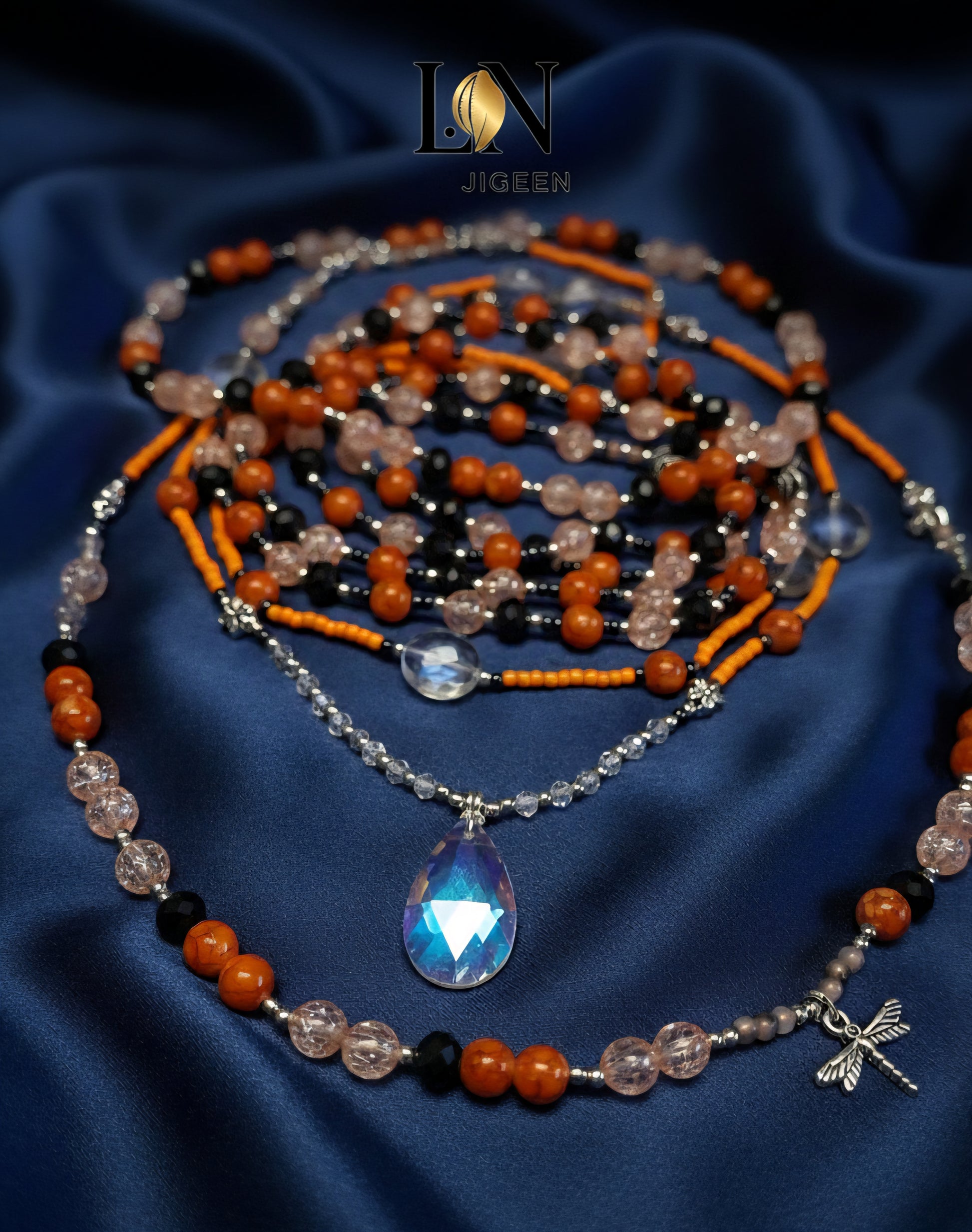 Gros plan professionnel des perles de reins Baya artisanal non portées, sur un fond de satin bleu marine. Détails des trois rangées de perles aux couleurs terre, rose poudré et noir, incluant un pendentif central. Logo de la marque affiché.