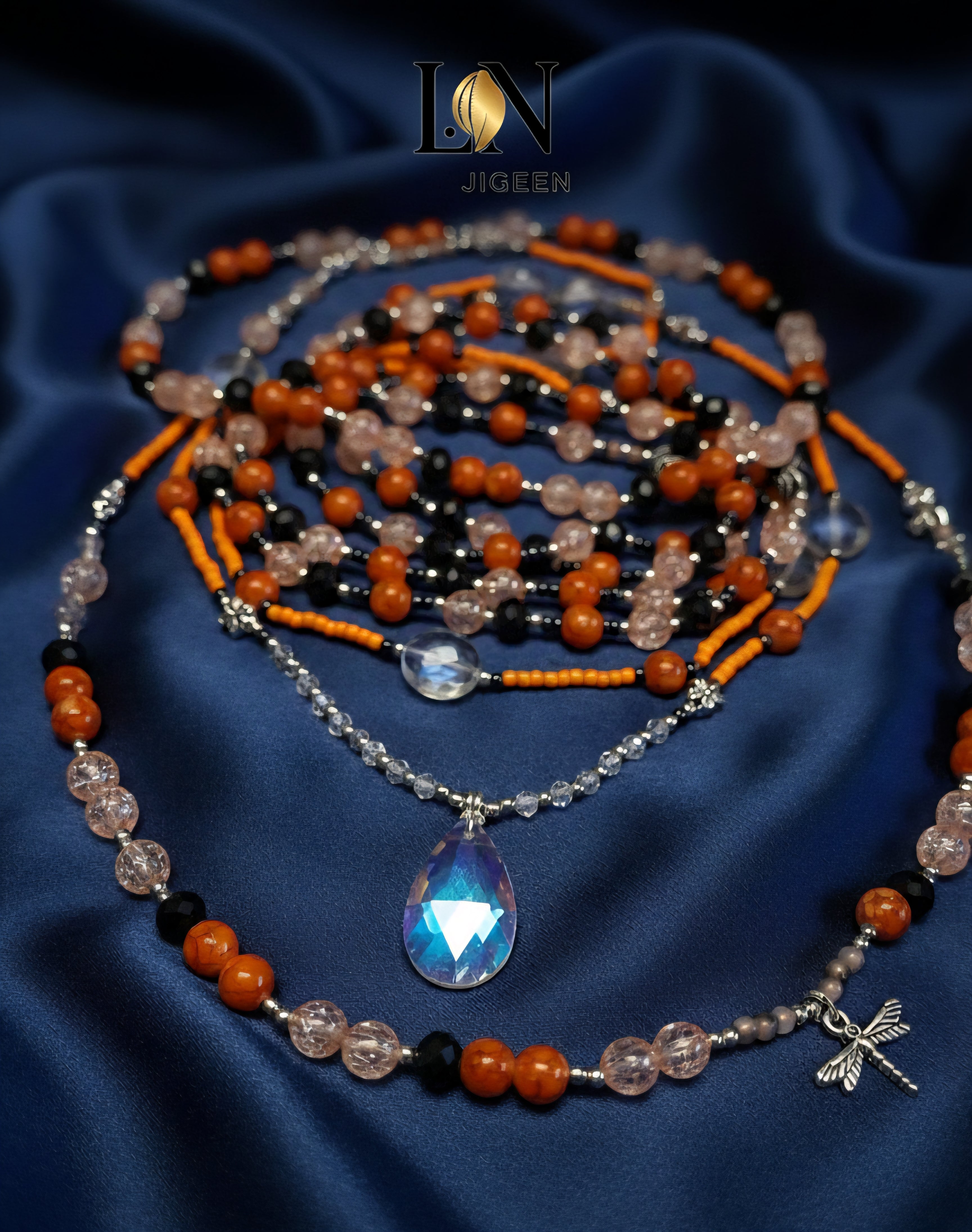 Gros plan professionnel des perles de reins Baya artisanal non portées, sur un fond de satin bleu marine. Détails des trois rangées de perles aux couleurs terre, rose poudré et noir, incluant un pendentif central. Logo de la marque affiché.