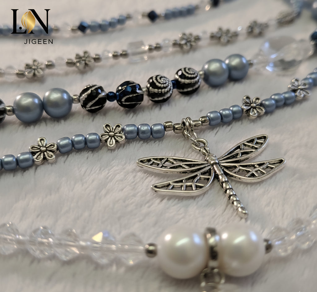 Gros plan sur les détails d'une perle de hanche multi-rangs : breloque libellule argentée, perles nacrées bleues et blanches, perles de cristal transparentes et noires, mettant en évidence les textures et finitions du bijou.