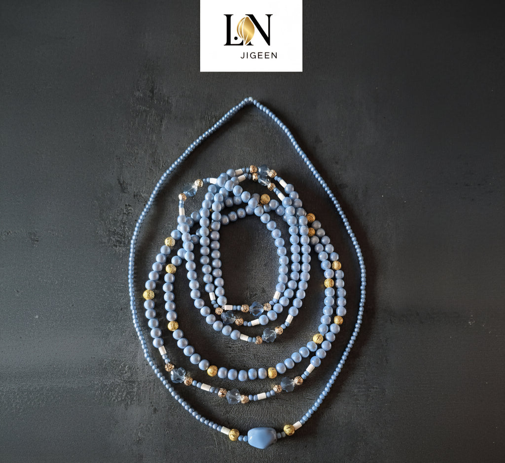 Perles de hanche (Waist Beads) 'LN JIGEEN' de couleur bleu pastel et or sur fond gris anthracite. Le bijou est composé de plusieurs rangs de perles rondes, de perles dorées texturées, de perles blanches et de perles de cristal bleu transparent. Le rang extérieur présente une perle centrale bleue de forme ovale