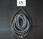 Perles de hanche (Waist Beads) 'LN JIGEEN' de couleur bleu pastel et or sur fond gris anthracite. Le bijou est composé de plusieurs rangs de perles rondes, de perles dorées texturées, de perles blanches et de perles de cristal bleu transparent. Le rang extérieur présente une perle centrale bleue de forme ovale