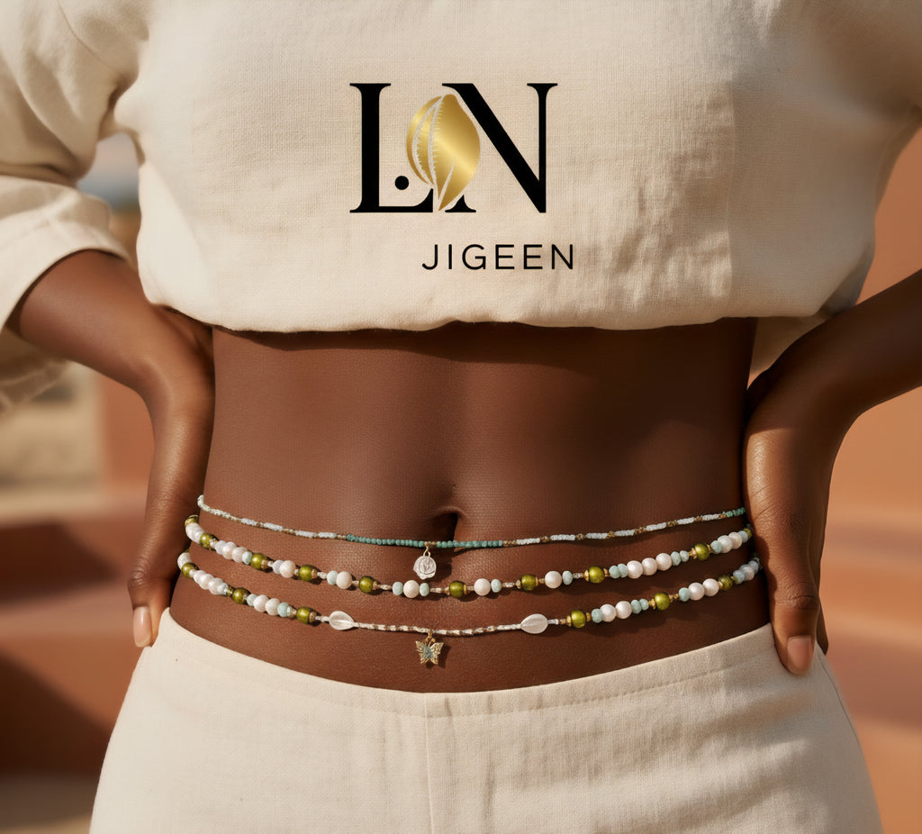 Gros plan professionnel sur des perles de hanche Bahia à trois rangées (vert sauge, blanc nacré, or) portées sur le ventre d'une femme à la peau foncée, vêtue de lin beige. Les perles de taille sont ajustées et arborent un pendentif papillon. Image de marketing pour bijoux culturels africains.