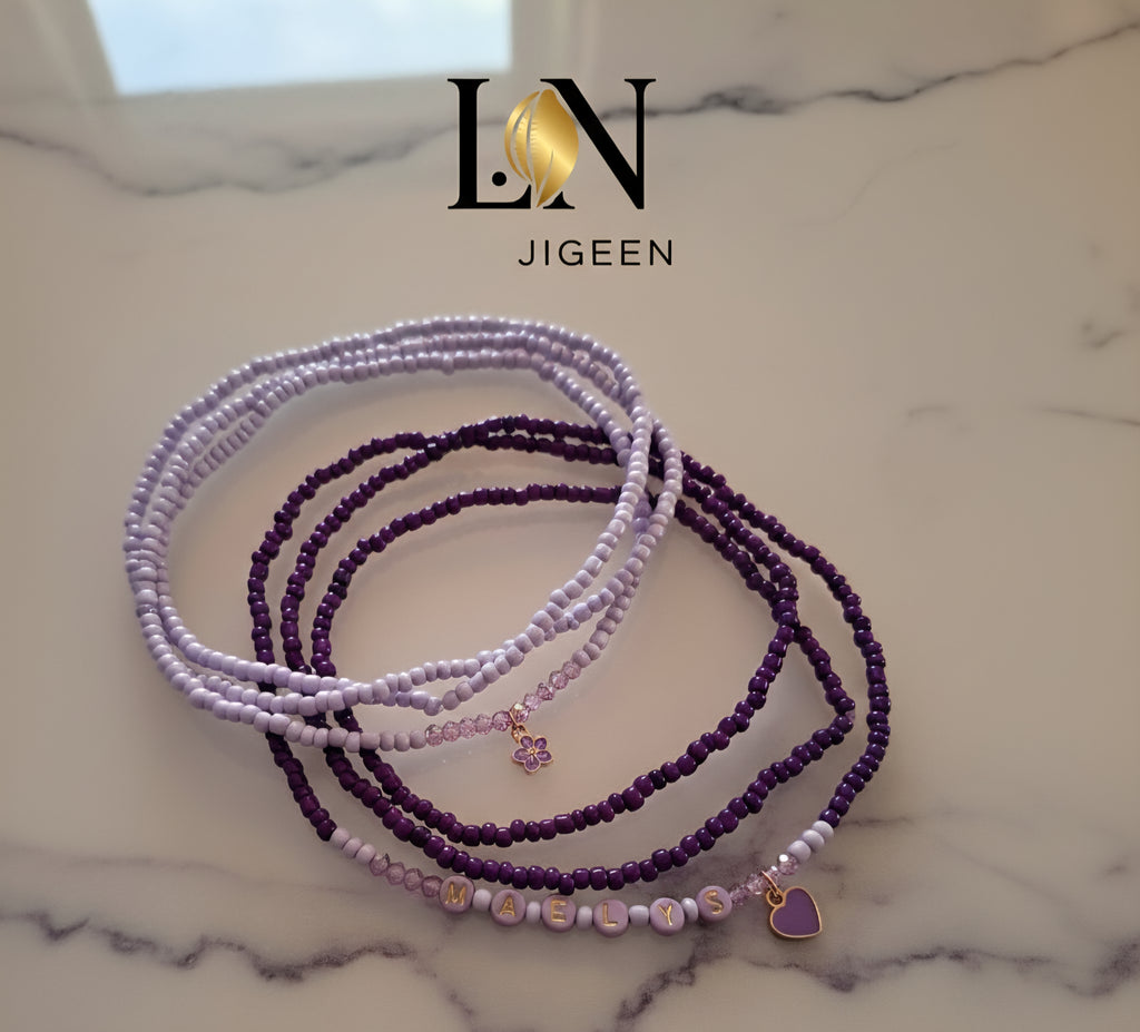 Quatre rangs de perles de reins (bijoux de taille) violettes et mauves, posées sur une surface en marbre clair. Les perles de couleur lavande sont plus fines, et un des rangs plus foncés comporte des perles formant le mot 'MAELYS' ainsi qu'un petit pendentif en forme de cœur violet. Le logo 'LN JIGEEN' est positionné en haut de l'image.