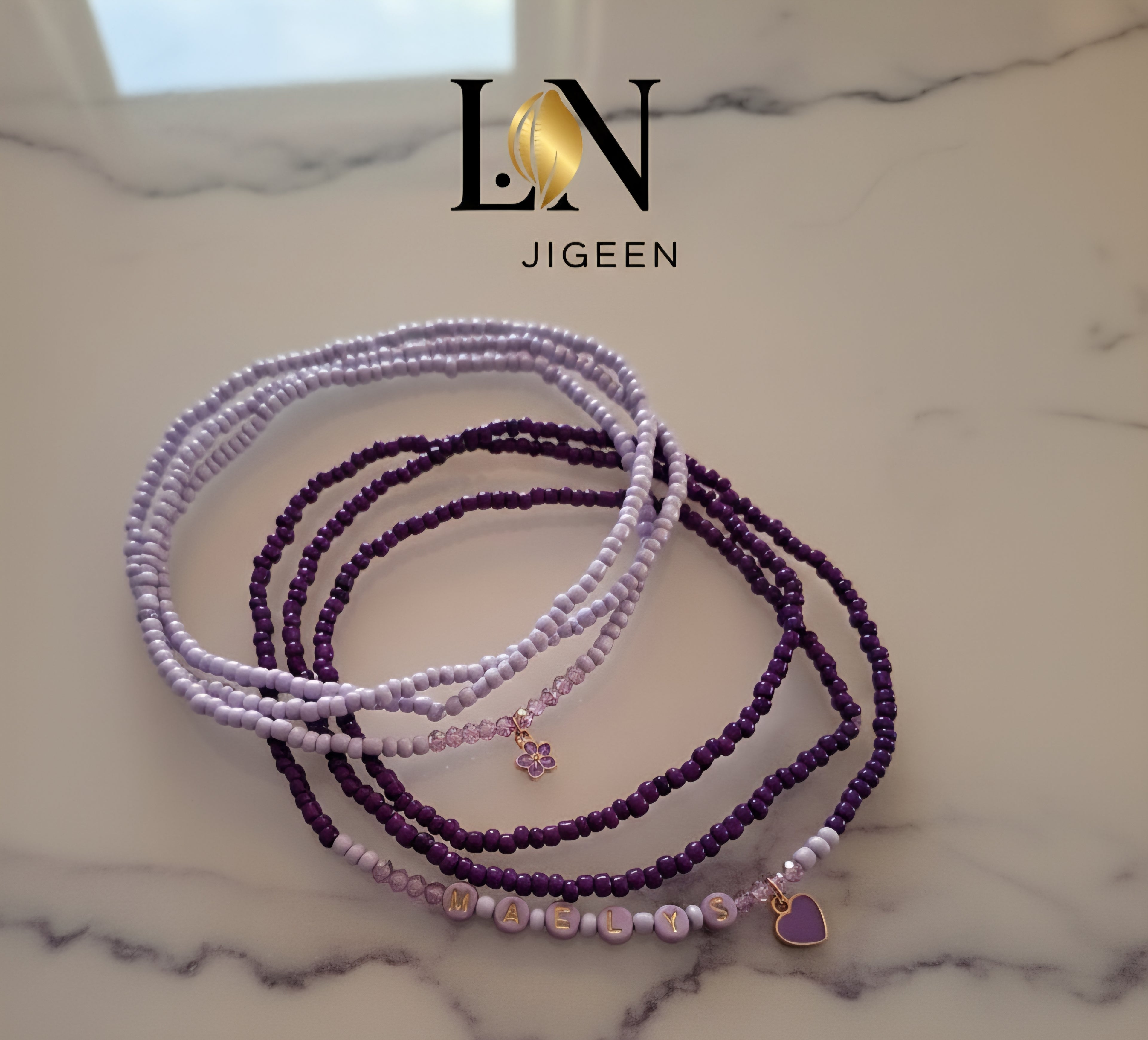 Quatre rangs de perles de reins (bijoux de taille) violettes et mauves, posées sur une surface en marbre clair. Les perles de couleur lavande sont plus fines, et un des rangs plus foncés comporte des perles formant le mot 'MAELYS' ainsi qu'un petit pendentif en forme de cœur violet. Le logo 'LN JIGEEN' est positionné en haut de l'image.
